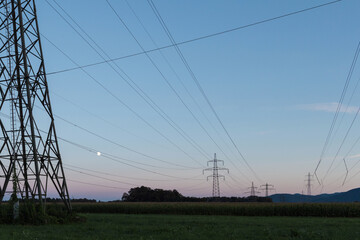 Obraz premium Power lines and transmission towers run across fields as the moon rises in the sky, showing energy development and electricity needs.
