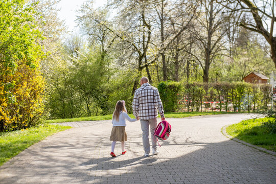 Grandfather walking hand in hand with granddaughter across sunny park, little girl wearing a pink school backpack, Walking to school spring day, education concept, family bonding, outdoor lifestyle