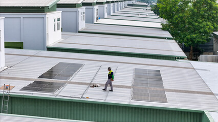 technician in safety gear inspects solar panels on a metal rooftop, holding a tool. The scene emphasizes renewable energy, solar technology, and maintenance of sustainable infrastructure. © ultramansk