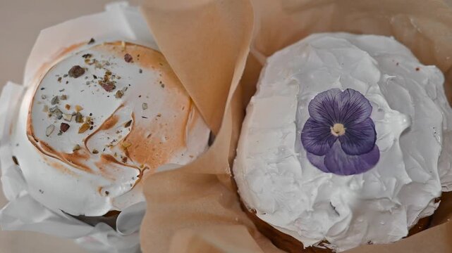Cakes are placed on a neutral surface with a plain background. Each cake features decorative frosting and a purple flower on top, showcasing a focus on simple presentation