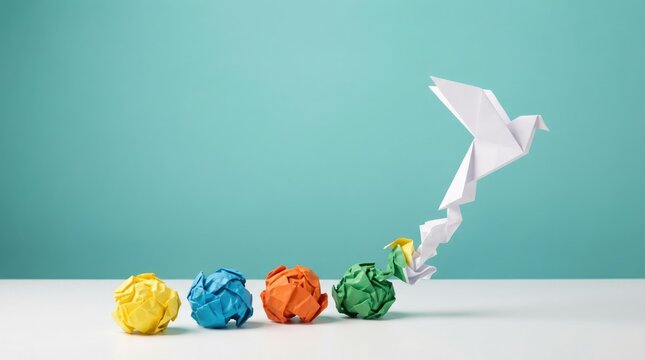 White origami bird flying away from a row of colorful crumpled paper balls on a white surface against a solid teal background