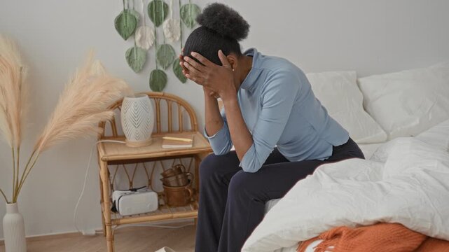 Woman seated on bed covering her face with hands while leaning forward in a styled bedroom studio with bedding and nightstand details; anxiety.