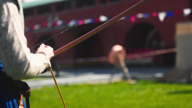 Medieval archery tournament in a castle yard with a woman shooting an arrow from a wooden bow in slow motion. Female archer in traditional medieval dress during historical reenactment, outdoor scene w