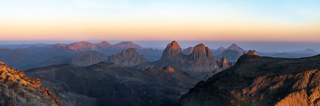 Elevated sunset view from Assekrem summit of the Atakor mountain chain. Jagged volcanic peaks glow orange against a soft gradient colorful sky in the Hoggar plateau of Tamanrasset, Algeria.