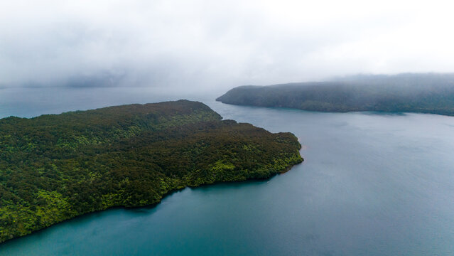 Aerial view of dark waters embrace land masses cloaked in deep green foliage under a blanket of heavy mist, Manapouri, New Zealand.