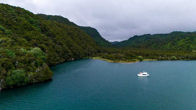 Aerial view of a serene lake reflecting the overcast sky, with a single boat amidst the lush, green, forested hills, Lake Tarawera, New Zealand.