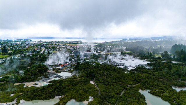 Aerial view of geothermal activity steaming through the landscape, contrasting with the lush greenery and the distant lake under a cloudy sky, Rotorua, New Zealand.