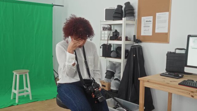 Woman photographer covering her face with hand while holding a dslr camera in a studio with green screen and equipment; creative frustration.