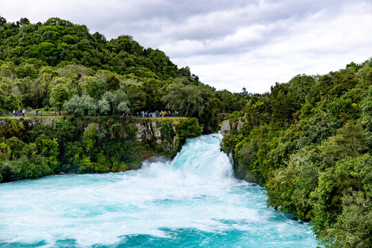 View of powerful turquoise waters thunder through a narrow gorge, framed by lush green forests under a cloudy sky, Huka Falls, Waikato Region, New Zealand.