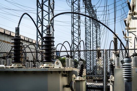 Large electrical substation with high-voltage equipment under bright blue sky. Black insulators and curved conductors dominate the scene. Metal towers stretch upward, carrying power lines