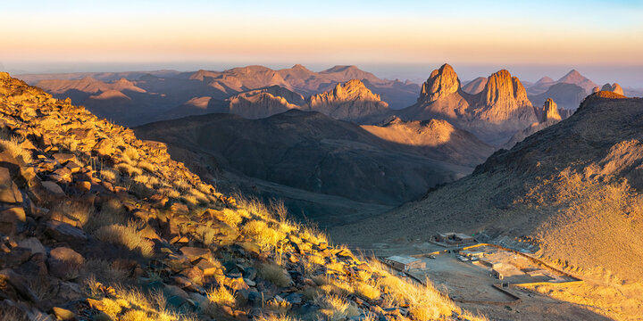 Elevated sunset view from Assekrem summit of the Atakor mountain chain. Jagged volcanic peaks glow orange against a soft gradient colorful sky in the Hoggar plateau of Tamanrasset, Algeria.