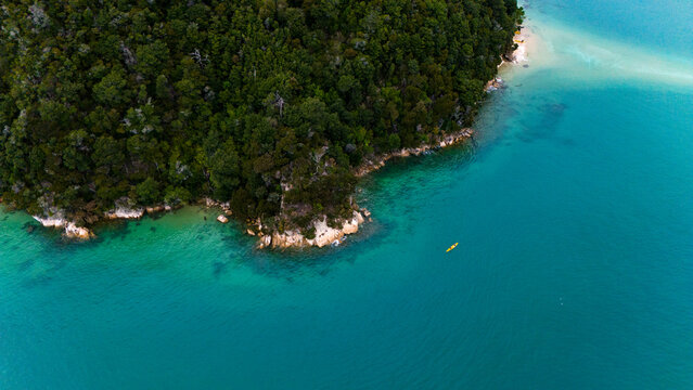 Aerial view of verdant forest meeting the turquoise sea, a lone kayak adrift in the vast expanse, Abel Tasman National Park, Tasman Region, New Zealand.