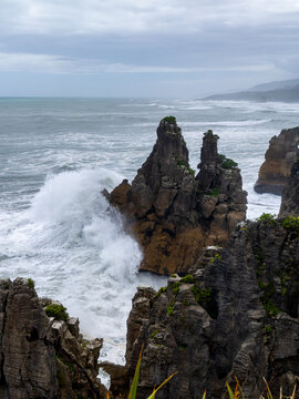 View of powerful waves crashing against the rugged, dark rock formations under an overcast sky, creating a dramatic coastal scene, Pancake Rocks, West Coast Region, New Zealand.