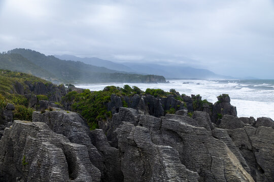 View of rugged, layered pancake rocks meet the turbulent, foamy surf under a grey, brooding sky, Pancake Rocks and Blowholes Walk, West Coast Region, New Zealand.