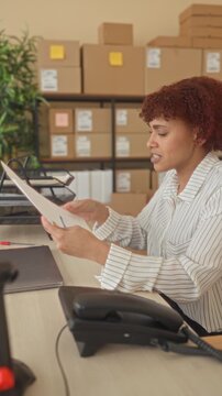 Woman reading a document and holding paper while touching her forehead amid stacked cardboard boxes and parcels at an office building, phone on desk; stressed focus.