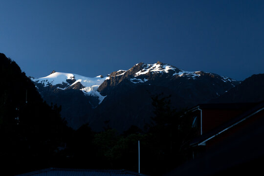 View of snow-capped mountains gleaming under a bright sky, casting long shadows over the landscape, a serene vista, Franz Josef Glacier, West Coast Region, New Zealand.