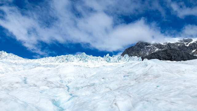 View of the glacier's icy expanse meets a rugged mountain under a bright sky with fluffy clouds, creating a striking contrast, Franz Josef, West Coast Region, New Zealand.