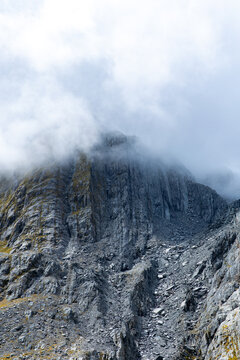 View of a rugged mountain range shrouded in ethereal mist, with textured rocks and sparse vegetation, showcasing nature's grandeur, Franz Josef, West Coast Region, New Zealand.