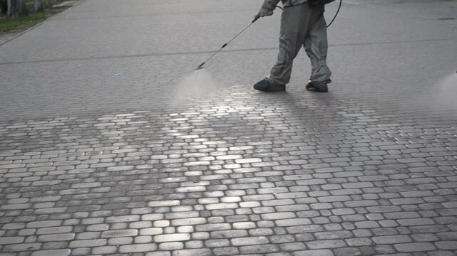 Man in airtight suit sprays disinfecting liquid chemicals antiseptic on street in city on day. Sanitation workers clean pavement, crosswalk. Sanitary measures in public place. Coronavirus pandemic