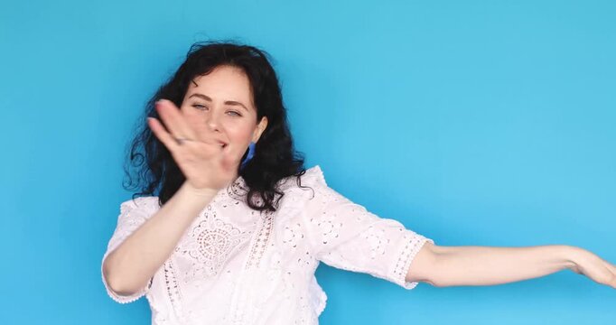 Happy young woman with curly hair singing and dancing in studio, joyful girl in white lace blouse lip-syncing and moving gracefully against blue background, positive emotions and music lifestyle.