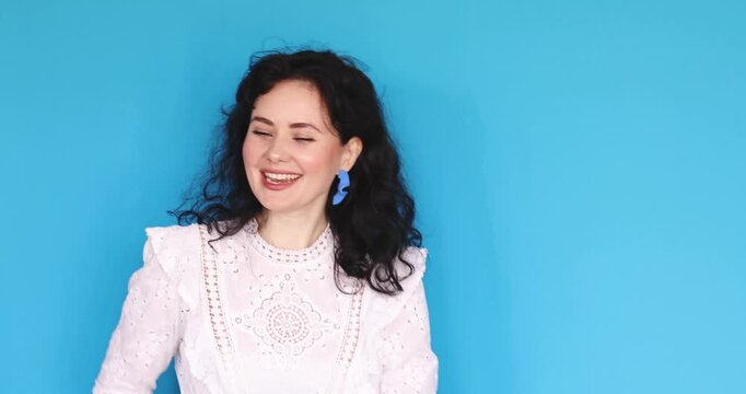 Cheerful young woman with curly hair singing and dancing against blue studio background, happy portrait of girl in white lace blouse lip-syncing and expressing joy and positivity.