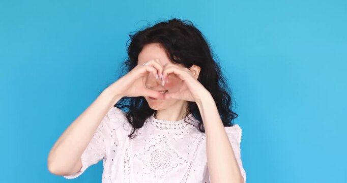 Playful woman curly hair dancing, winking and making heart gesture in studio, joyful girl in white blouse lip-syncing and showing love, flirting gracefully against blue background, positive emotions.