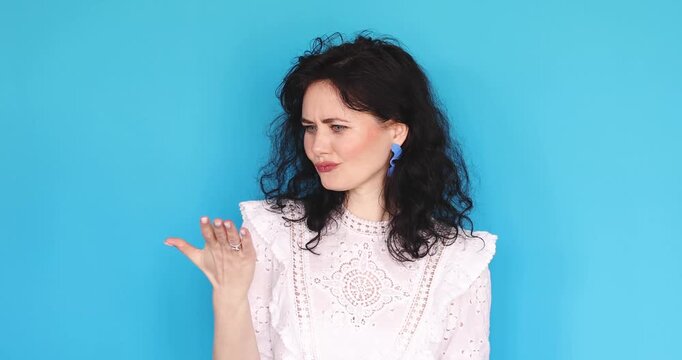 Young curly woman showing money gesture with fingers, asking for payment or cash, looking skeptical and demanding with expressive face on blue background, brunette lady making pay me sign