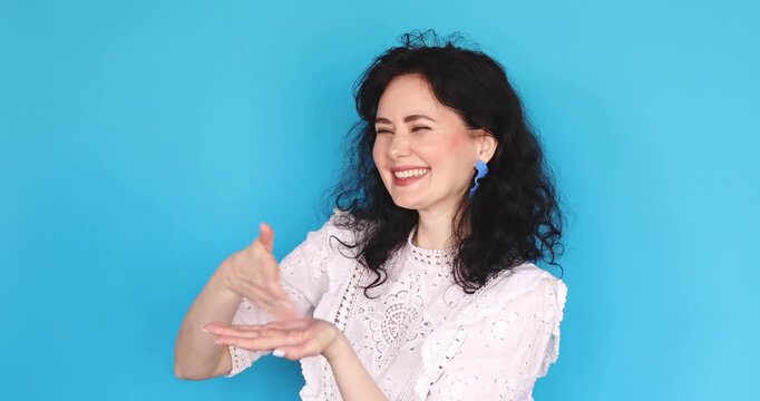 Excited young woman with curly hair in white lace top throwing imaginary money, scattering invisible bills with expressive hand gestures and laughing joyfully on blue background