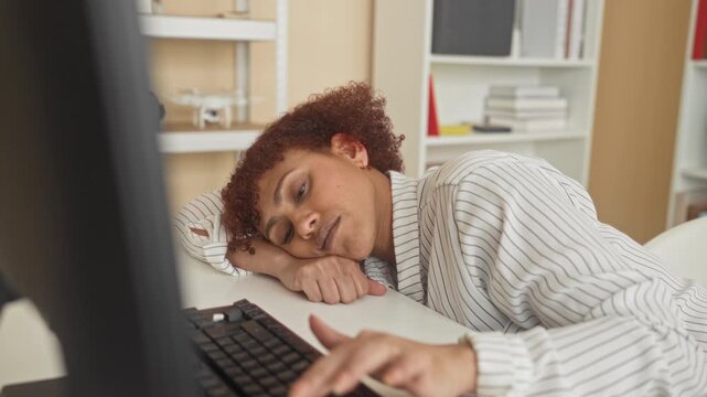 Woman resting head on desk and reaching for keyboard beside monitor in office building, leaning forward with slumped posture and blank expression; fatigue.