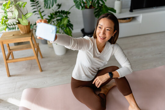 East Asian woman sitting on a yoga mat at home taking a selfie with her smartphone. Bright, cheerful wellness moment with indoor plants, fitness routine, and positive, relaxed lifestyle