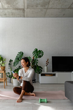 East Asian woman sitting on a yoga mat at home, using her smartphone after a workout. Calm, focused moment with indoor plants and soft daylight, reflecting a balanced, modern wellness lifestyle