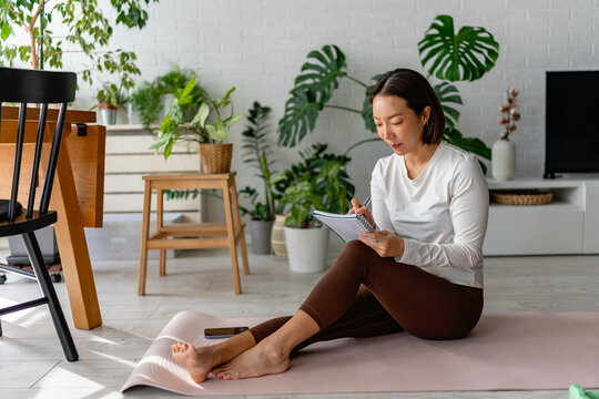 East Asian woman sitting on a yoga mat at home, writing notes in a notebook after a workout. Calm, focused wellness moment with indoor plants and natural light in a cozy living space