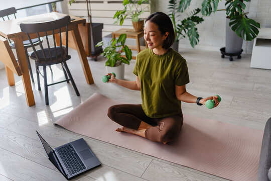Asian woman exercising at home on a yoga mat with dumbbells while following a laptop workout. Healthy lifestyle scene showing motivation, focus and active living indoors.