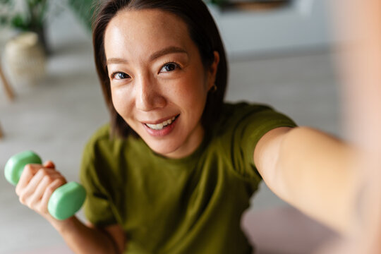 Smiling Asian woman taking a selfie while exercising with a dumbbell at home. Fitness lifestyle scene showing motivation, energy and positive mood during a home workout.