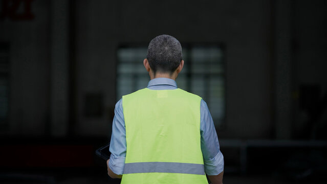 A middle-aged male worker wearing a fluorescent yellow safety vest viewed from behind in an industrial facility