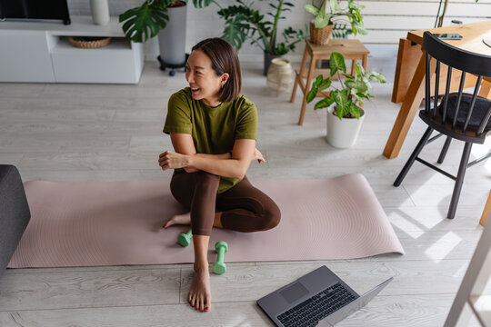 Asian woman resting on a yoga mat after a home workout, smiling and looking aside. Relaxed fitness lifestyle scene with dumbbells and laptop in a bright, plant-filled living space.