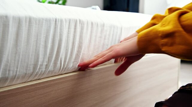 Person adjusts bed sheet on wooden bed frame in bright bedroom setting