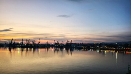 Fototapeta premium evening harbor panorama with glowing lights and cranes mirrored in water creating atmospheric urban waterfront scene with soft pastel sky tones