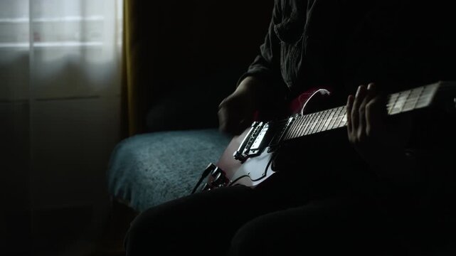 unrecognizable man in black denim playing electric red sg guitar in the day at home. low key light