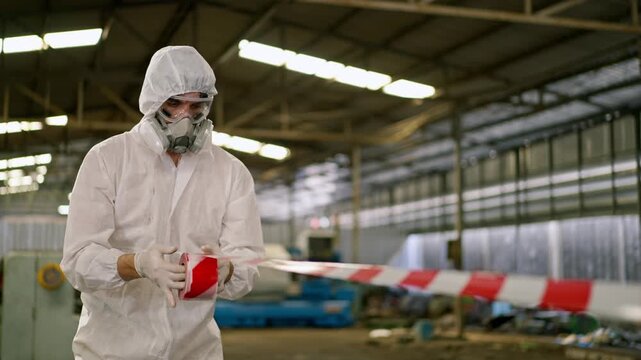 Hazmat suit respirator protective clothing factory safety worker sealing red and white caution tape in industrial warehouse during contamination biohazard barrier control for workplace protection