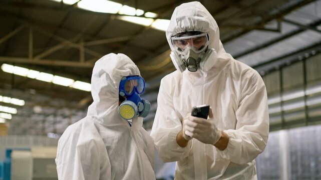 Worker hazmat suit respirator protective clothing factory smartphone safety inspection during industrial contamination checks by two factory worker in protective gear using smartphone inside factory