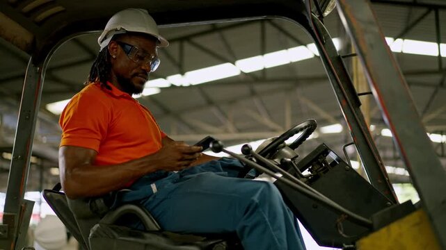 Forklift operator using smartphone inside warehouse cab during material handling, wearing hard hat and safety glasses, focused industrial worker logistics facility, seated forklift operator checking