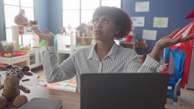 Woman teacher brunette with laptop resting chin on hand amid kindergarten toys in school building; weariness.