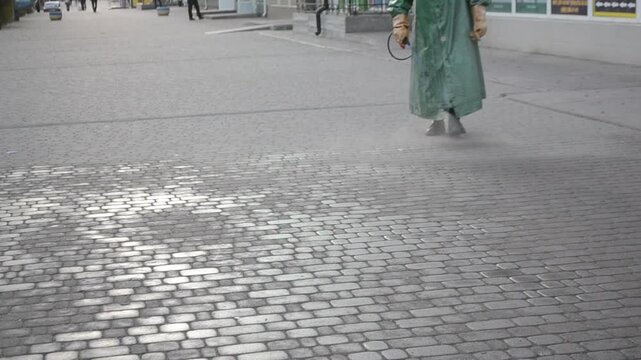 Man in airtight suit sprays disinfecting liquid chemicals antiseptic on street in city on day. Sanitation workers clean pavement, crosswalk. Sanitary measures in public place. Coronavirus pandemic