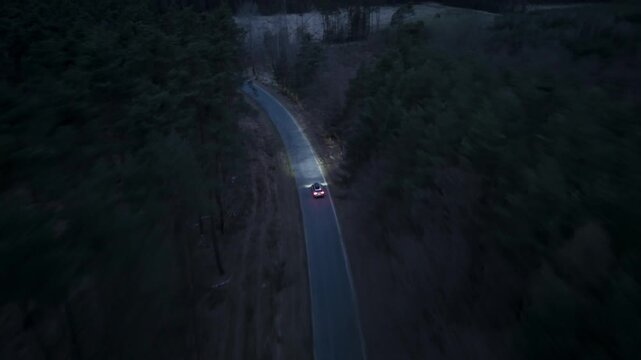 Aerial night shot of a car driving through dark forest with headlights on as drone follows vehicle along straight road