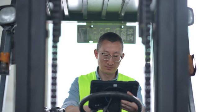 Middle-aged male forklift operator wearing safety vest and using tablet computer in warehouse