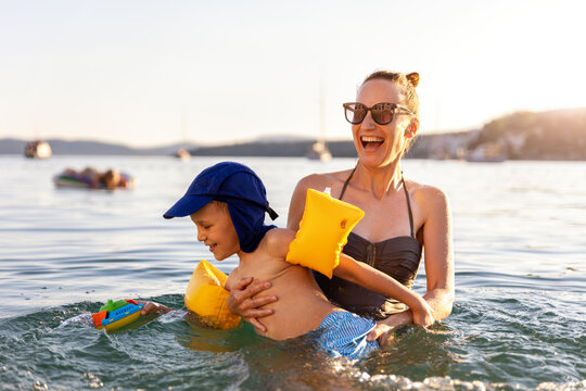 Mother laughing have fun hold child sea swim warm summer sunset. Joyful family vacation candid moment expression playful kid wear armbands splashing water calm seaside coastline travel destination