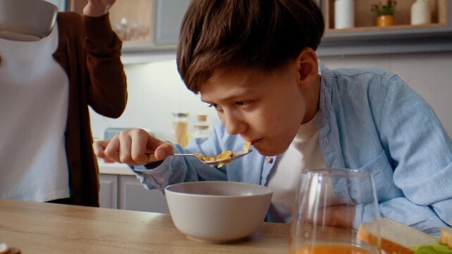 Preteen boy eats corn flakes with milk and nods head to mother