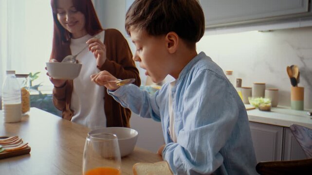 Happy little boy eats corn flakes with milk by mother in kitchen