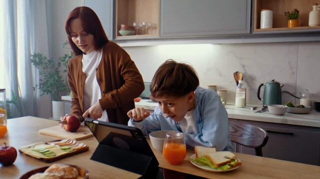 Woman cuts apple as preteen son eats breakfast and watches video
